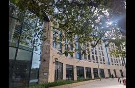 A beige brickwork building with a number of windows and doors. The building is partly covered by a large leafy tree
